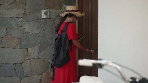 Side view serious young woman closing door walking to bicycle leaving in slow motion. Portrait of Caucasian millennial lady in straw hat sunglasses and red dress cycling on sunny summer day outdoors