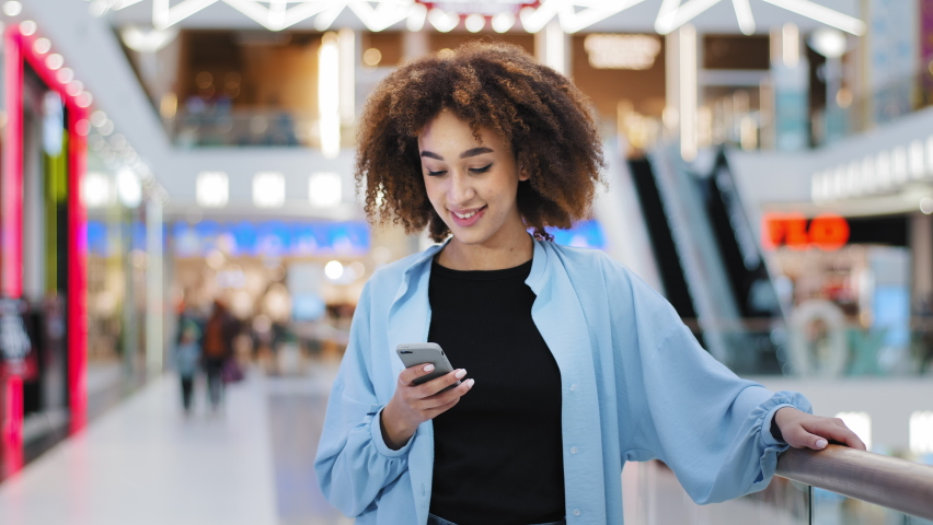 Young girl african american woman customer blogger girlfriend standing in shopping mall looking at mobile phone chatting browsing reading message waiting date using smartphone app for online shopping
