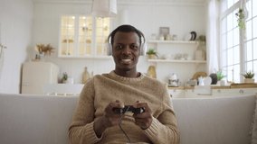 Young African American man wins playing video game console in kitchen. Happy player in headphones screams and laughs at home close view