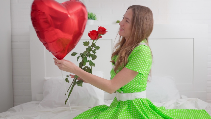 A beautiful girl in a green dress with gifts of a bouquet of red roses and a balloon in the shape of a heart is sitting on a bed on a white bedspread. Valentine's day. Relationships. Love. March 8 Royalty-Free Stock Footage #1090365869