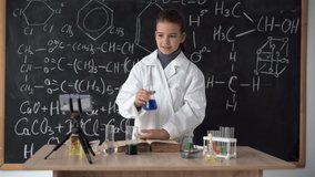 ittle scientist girl in white coat leads an online video blog, records video using smartphone. Research and education. Chemical laboratory with glass flasks. Schoolgirl conducts a chemical experiment