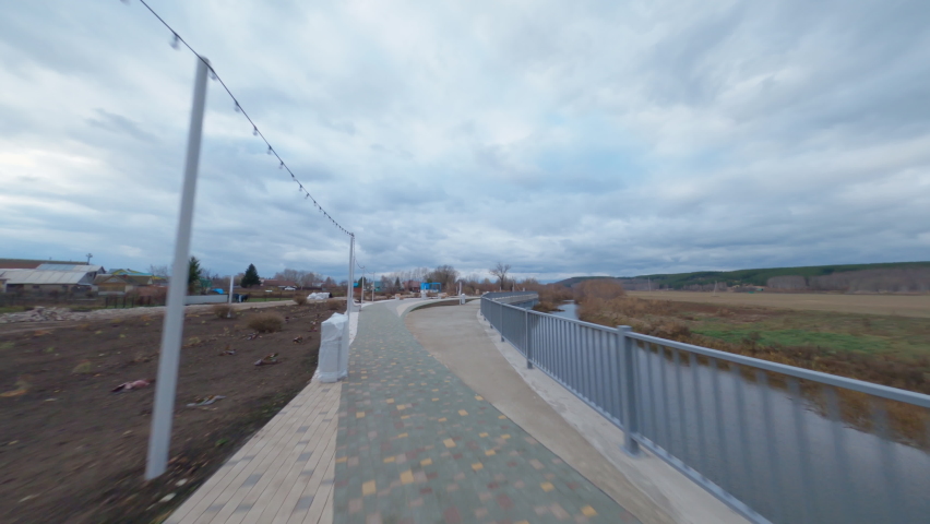 Paved pathway with railing near narrow canal in young city park under sky with heavy clouds aerial first point view