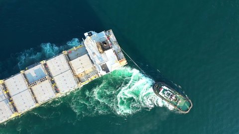 Aerial drone top down video of tow - tug boat assisting by pulling empty container ship to depart from container terminal port