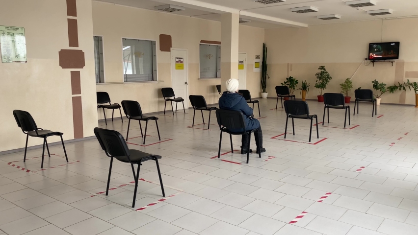 Concept of social distancing in waiting room at station. One masked man is sitting on chair, keeping his distance. Woman leaves leaving an empty hall. New normal life during pandemic.
