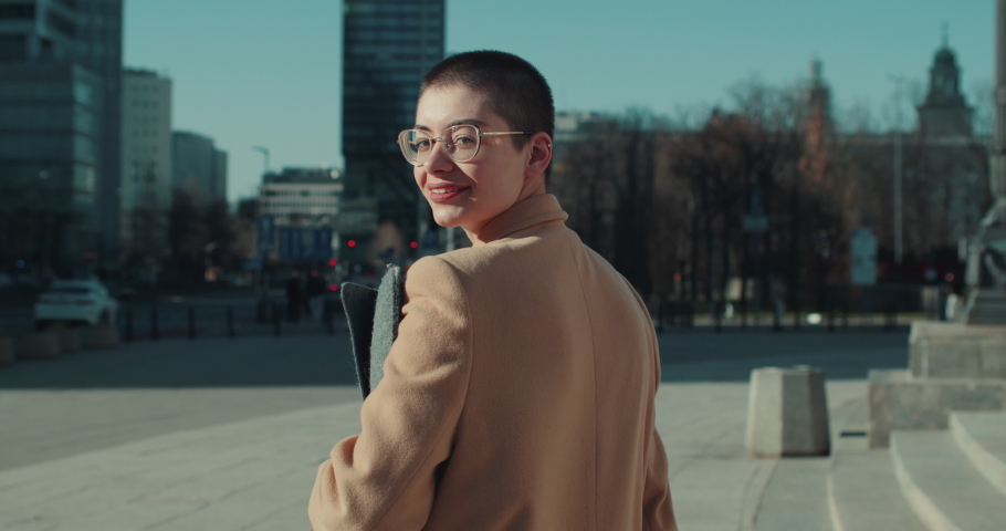 Positive young 20s smiling business woman with short hair in eyeglasses walks outside looking back at camera slow motion Royalty-Free Stock Footage #1090385221