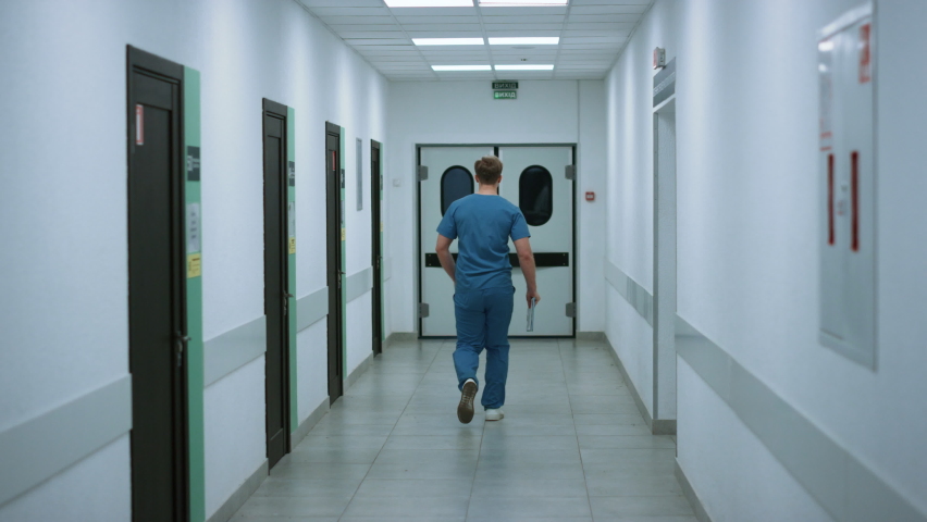 Unknown surgeon going to operation room holding folder back view. Young doctor walking on hospital corridor wearing blue uniform. Medical professional rushing to help patient down clinic hallway. | Shutterstock HD Video #1090387149