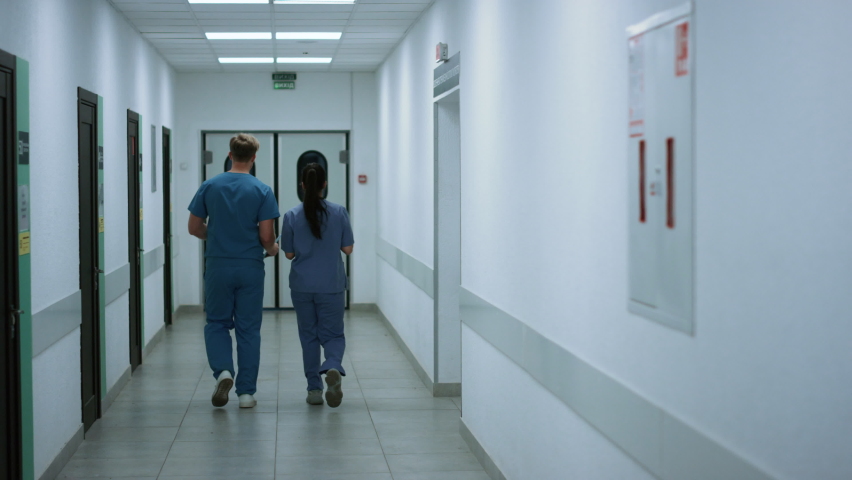 Unknown doctor with nurse moving away down hallway back view. Man surgeon consulting woman colleague walking in hospital corridor. Medical professionals discussing health care in clinic hall. | Shutterstock HD Video #1090387153