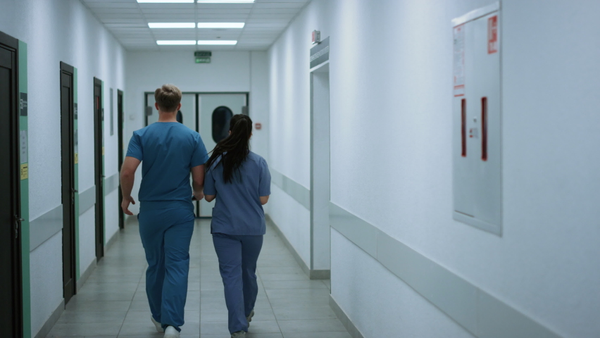 Doctors colleagues walking down clinic corridor talking about work. Back view two medics going to emergency room door. Surgeon man consulting woman physician about patient in hospital hallway.  | Shutterstock HD Video #1090387163