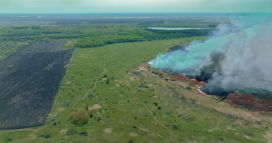 Copter flies over the fire in the village near the river, the grass is on fire. Black smoke and tubers rise into the sky, an ecological disaster. Natural disaster, burning field near the water. 4k