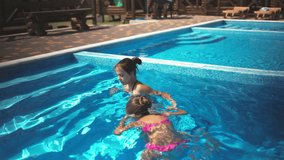 An older sister in a bright leopard swimsuit, pulls out and scolds her younger sister who does not want to climb out of a large deep pool with clear blue transparent water