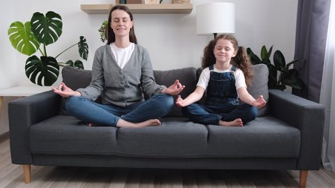 Doing like mommy. Calm young mother and little daughter having fun sit cross legged on comfy couch at modern studio flat do yoga together. Funny small kid girl imitates mom in meditation practice
