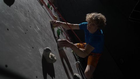 Cinematic lighting, young man professional climber training on a climbing wall, practicing rock-climbing and moving up, using insurance.
