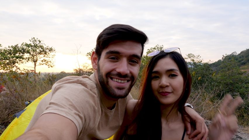 Happy love couple take photo and selfie during camping and relax in forest, beautiful asian woman and caucasian man happiness spend time tpgether in front of tent in evening.