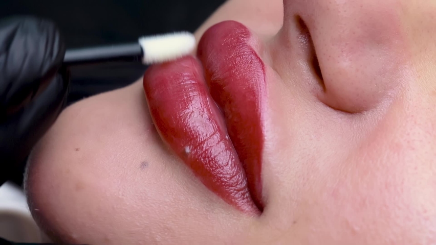 A close-up of the model's lips is made with permanent red makeup, the master runs a cotton swab over the model's lips, applying a moisturizing gel to the lips | Shutterstock HD Video #1090396565