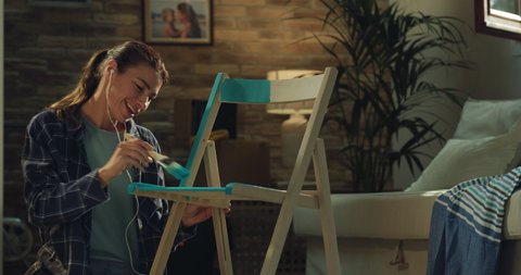 Cinematic shot of young creative woman is applying paint with paintbrush on wooden chair to remodeling and renovation house by herself with diy techniques at home.