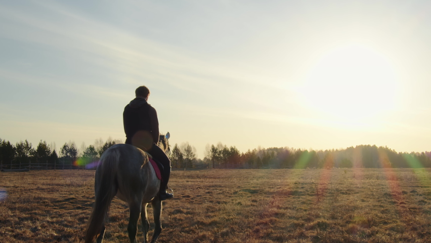 A large man slowly rides a horse across the field. He is calm and peaceful. Away forest. There is a blue sky above his head. Only their silhouettes are visible from behind the sun Royalty-Free Stock Footage #1090400631