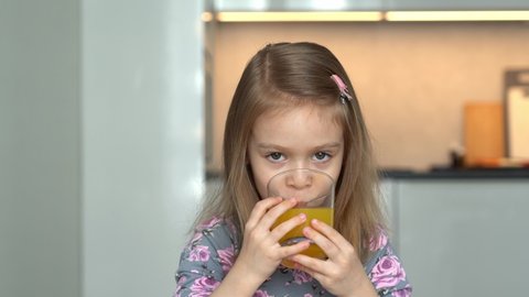 Little girl looks at camera, smiles and drinks orange juice. Happy child drinks juice on kitchen background