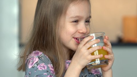 Close up of little girl laughs and fun drinks orange juice from glass. Happy child drinks juice on kitchen at home