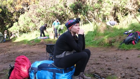 A hiker with action cams on her head sits on bags and waits for the group to gather and pack for the hike while climbing Mount Kilimanjaro at Machame camp