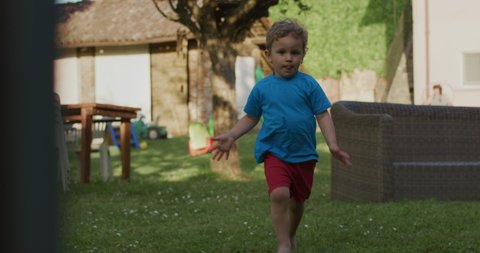 Cinematic authentic shot of happy smiling toddler baby boy is running on green lawn while having fun playing outside his house.