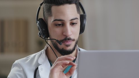 Professional male arab young doctor medical assistant in medical coat wear headset with microphone make conference call on laptop use distance virtual teach computer app consult online in video chat