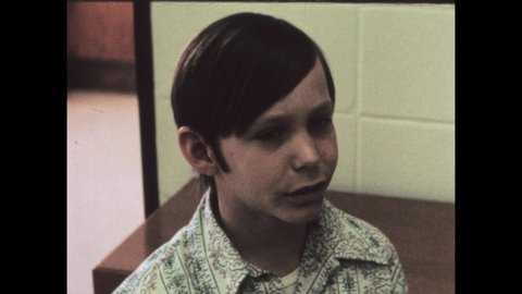 1970s: school boy with 1975 haircut and sideburns. Boy talks with man at school notice board. Boy brings projector to class.