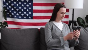 Smiling young American woman sitting on comfy sofa at home with USA flag waving hand using smartphone app enjoying online virtual chat video call with friends. 4 of July, Independence day celebration