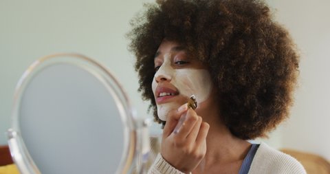 Happy biracial woman applying beauty face mask with brush. spending quality time at home alone.