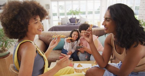 Happy diverse female friends sitting on sofa in living room, eating pizza. spending quality time at home.