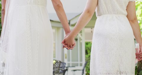 Happy diverse female couple holding hands and bunch of flowers in garden. wedding day and celebration concept.