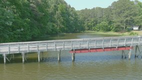  stunning aerial footage of the rippling green waters of the lake with a brown wooden bridge and dock on the water with lush green trees, grass and plants at Cauble Park at Lake Acworth in Acworth