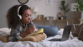 Young woman is studying in front of laptop and writing while lying on bed at home spbd. Close view of american african female looks at computer and listens to webinar, takes notes in notebook and lies