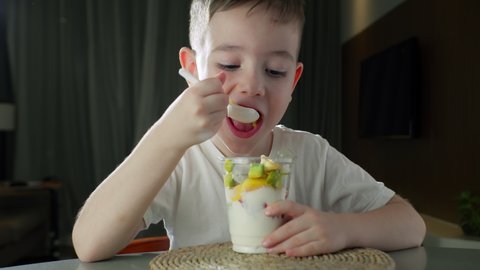 Child eats milk yogurt with tropical fruits. Healthy eating little boy dinner concept of childhood dream. Hungry daughter girl in the kitchen eats yogurt milk with fruits. Positive child smiling