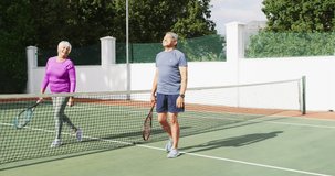 Video of happy biracial senior couple walking with rackets on tennis court. active retirement lifestyle, senior relationship and tennis training concept.