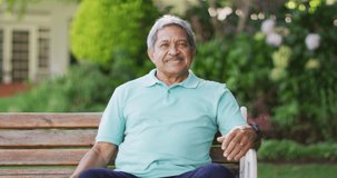 Video of happy biracial senior man sitting on bench in garden. active retirement lifestyle and spending time outdoors.
