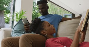 Video of happy african american couple lying on sofa with smartphone and book. love, relationship, togetherness and spending time with technology at home.
