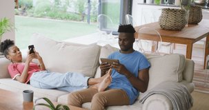 Video of happy african american couple sitting on sofa and using smartphone. love, relationship, togetherness and spending time with technology at home.