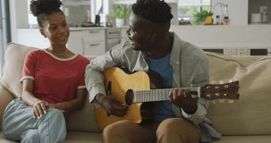 Video of happy african american couple sitting on sofa, singing and playing guitar. love, relationship, togetherness and spending quality time at home.