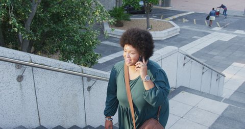 Happy plus size biracial woman walking on stairs and talking on smartphone in city. digital nomad on the go, body inclusivity.