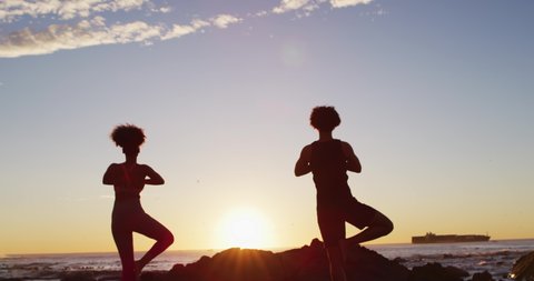 Rear view of african american couple practicing yoga together on rocks near the sea during sunset. love and relationship concept