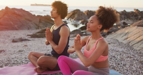 African american couple practicing yoga and meditating together on rocks near the sea during sunset. love and relationship concept