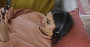 Video of happy biracial woman lying on sofa with headphones and using smartphone. leisure, relax, spending free time with technology at home concept.