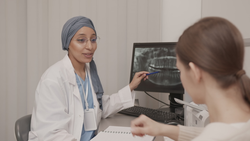 Medium slowmo of young Biracial female dentist talking to patient and pointing and teeth xray on computer monitor, planning further treatment at modern dental clinic | Shutterstock HD Video #1090432057