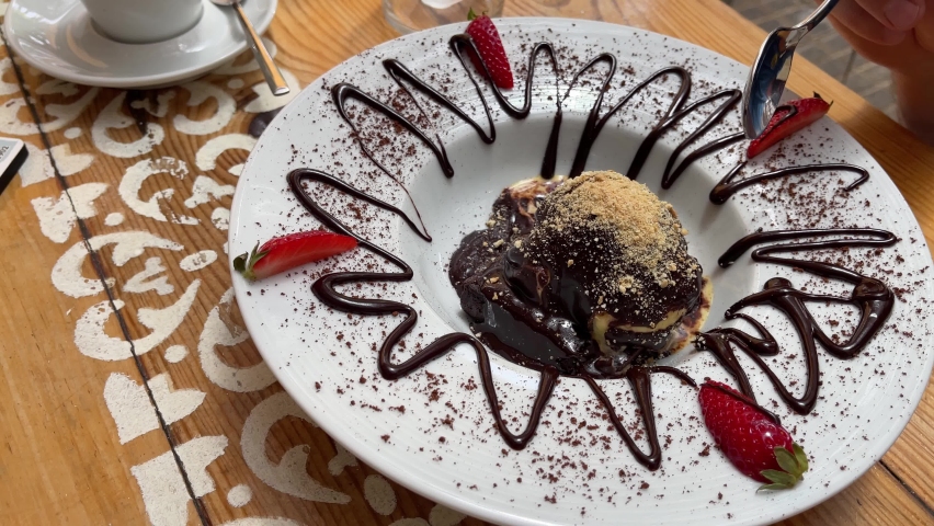 Spoon taking bite of chocolate brownie ice cream ball with fancy decoration of strawberries, syrup and powder on white plate. Succulent sweet dessert concept