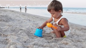 A little cheerful, enthusiastic boy plays with his toys on a sea sandy beach, building beads and turrets, smiling at someone behind the scenes on a bright sunny summer vacation