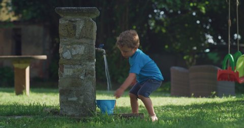 Cinematic authentic shot of carefree happy little toddler boy is having fun to play with fresh cool water from fountain in garden of his house in hot summer sunny day.