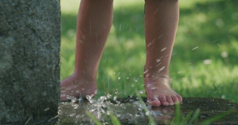 Cinematic authentic close up shot of water flows and splashes to little toddler boy feet while he having fun to play with fresh cool water from fountain in garden of his house in ho summer sunny day.