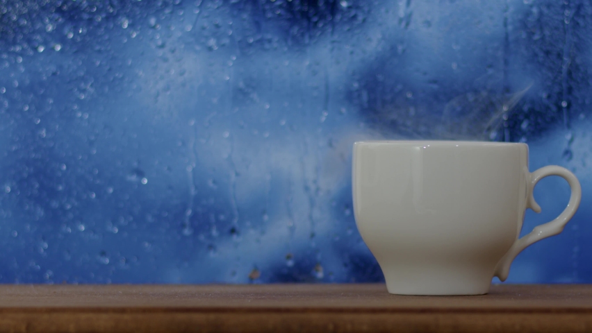 A glass of hot coffee in front of the window in rainy weather, raindrops drip on the window in beautiful weather. 4K. Royalty-Free Stock Footage #1090442281