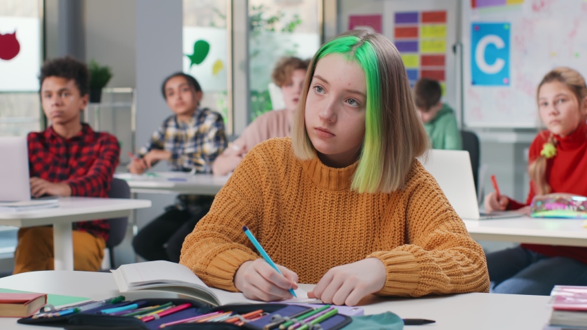 Teenage schoolgirl with green hair writing test in class in school. Portrait of hipster teen student doing homework sitting at desk in class Royalty-Free Stock Footage #1090442517