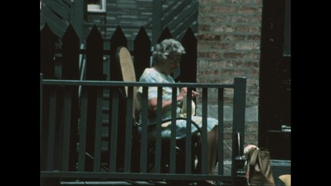 1970s: Woman sits on stoop in rocking chair, knits. Boy wears postal service uniform, pushes mail cart, waves, rubs chin. United States Post Office building.